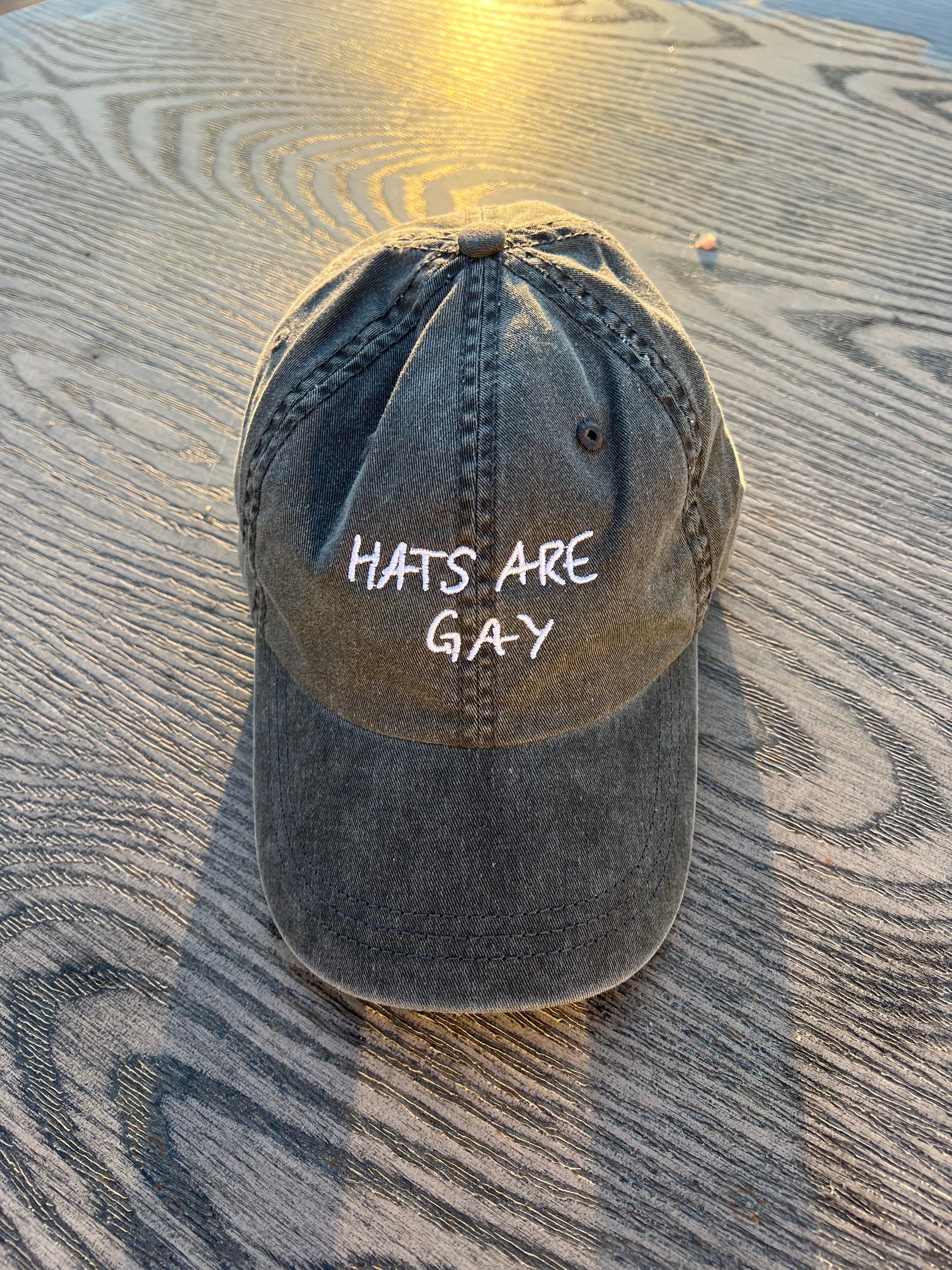 charcoal dad hat with the embroidered catchphrase "hats are gay" sitting on a wooden table to celebrate queer pride
