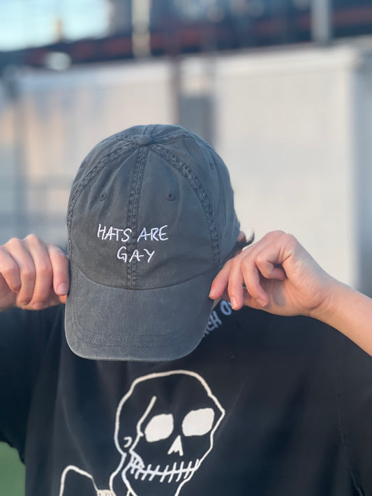 individual wearing a charcoal dad hat with their head tilted down with the embroidered catchphrase "hats are gay" to celebrate queer pride